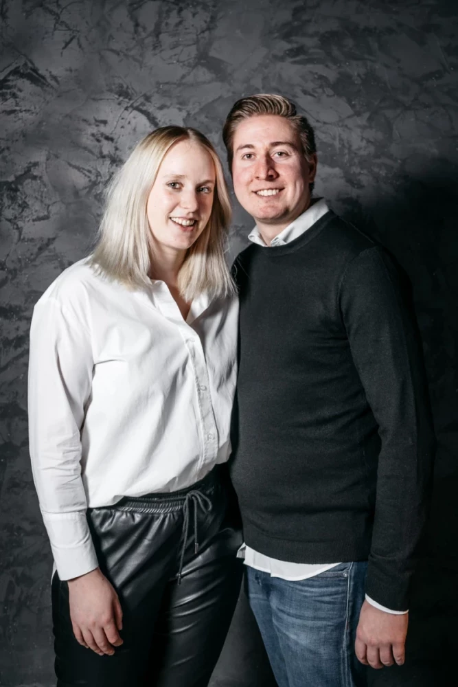 Zwei lächelnde Personen vor einer grauen Betonwand. Eine Frau in weißer Bluse und Lederhose steht links, ein Mann in schwarzem Pullover und blauer Jeans rechts von ihr. Beide stehen eng beieinander und wirken freundlich.
