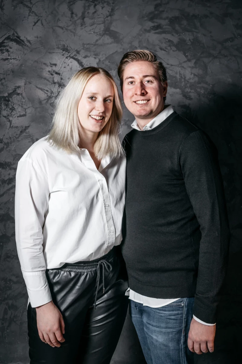 Zwei lächelnde Personen vor einer grauen Betonwand. Eine Frau in weißer Bluse und Lederhose steht links, ein Mann in schwarzem Pullover und blauer Jeans rechts von ihr. Beide stehen eng beieinander und wirken freundlich.