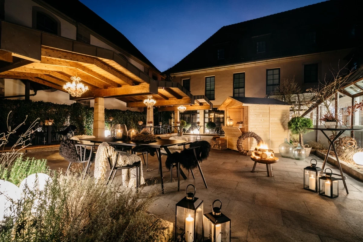 Abendliche Terrasse mit überdachtem Sitzbereich, warm beleuchtet von Laternen und Hängelampen. Gemütliche Atmosphäre mit Gartenmöbeln, Feuerstelle und dekorativen Laternen vor einer Hausfassade bei Nacht.
