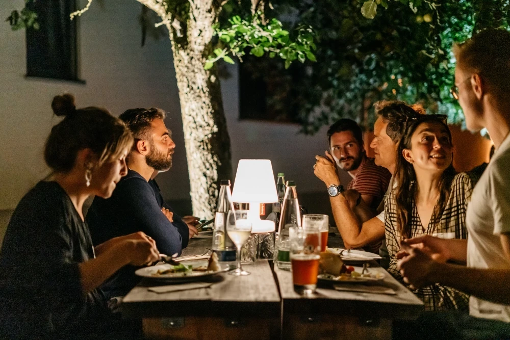 Mehrere Personen sitzen abends an einem Tisch im Freien, beleuchtet von einer Tischlampe. Sie unterhalten sich und essen gemeinsam, während im Hintergrund ein beleuchteter Baum steht.