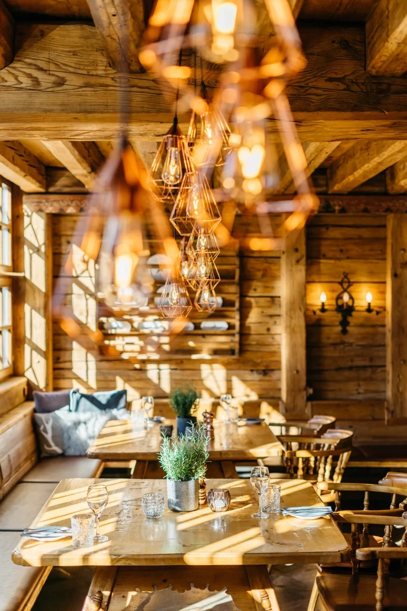Rustikal eingerichteter Gastraum mit Holzwänden und -balken, warm beleuchtet durch hängende Lampen und Wandleuchten. Gedeckte Holztische mit Blumen in Metallvasen, gemütliche Sitzecke mit Kissen und Sonnenlicht, das durch Holzfenster fällt.