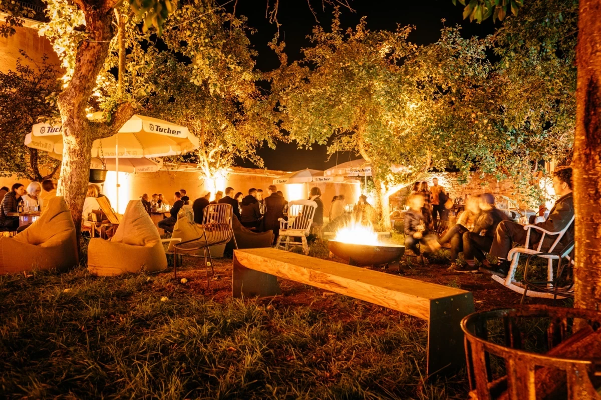 Abendliche Outdoorparty mit warmem Kerzenlicht, Bäumen und gemütlichen Sitzgelegenheiten. Menschen versammeln sich unter beleuchteten Pavillons und um ein gemütliches Lagerfeuer auf einer nächtlichen Wiese.