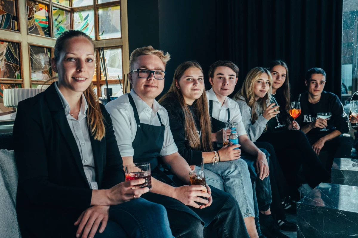 Acht junge Menschen sitzen nebeneinander in einem Café oder Restaurant und halten Getränke in der Hand. Sie tragen überwiegend dunklere Kleidung und lächeln in die Kamera. Die Gruppe wirkt locker und entspannt.