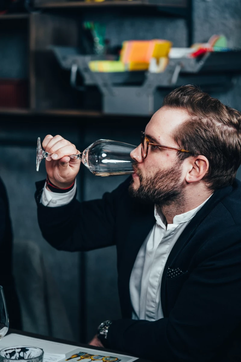 Mann mit Brille und schwarzem Sakko trinkt Wein aus einem Weinglas in einer dunklen Umgebenheit, konzentriert und professionell wirkend.