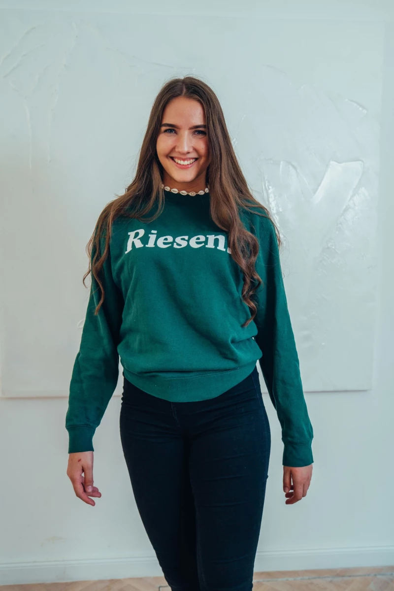 Junge Frau in grünem Pullover mit "Riesen"-Aufdruck und schwarzer Hose, die vor einer hellen Wand steht und lächelnd in die Kamera blickt.