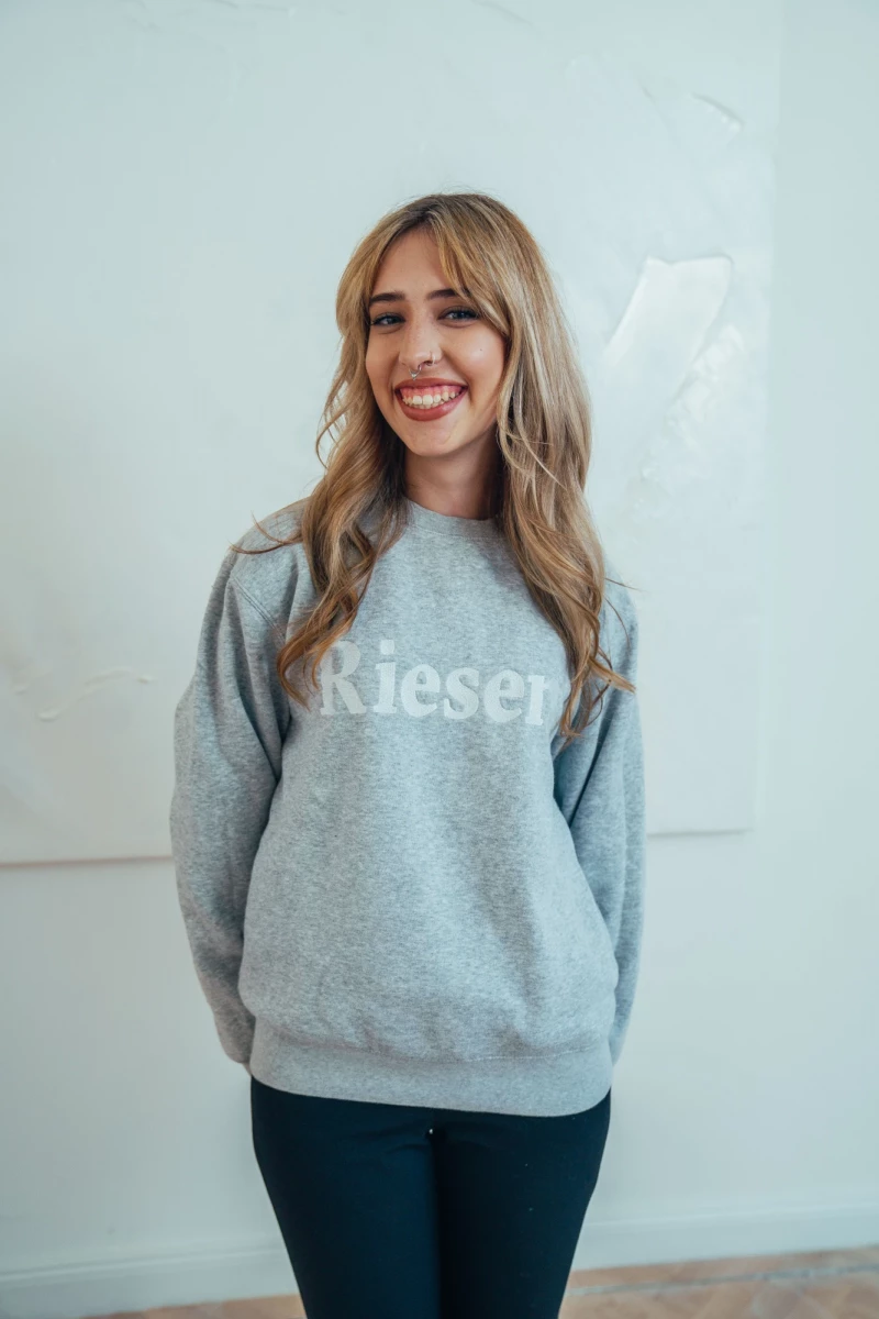 Eine lächelnde Frau mit welligen Haaren trägt einen grauen Pullover mit Riesengebirge-Schriftzug und dunkle Hose. Sie steht vor einer hellen Wand und lächelt fröhlich in die Kamera.