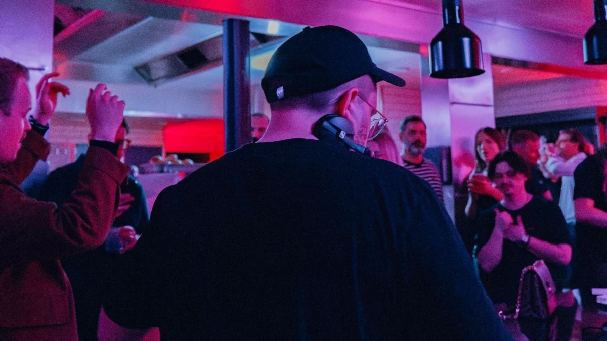 DJ mit schwarzer Kappe und Kopfh&ouml;rern steht vor einer tanzenden Menschenmenge in einem Raum mit roter und blauer Beleuchtung