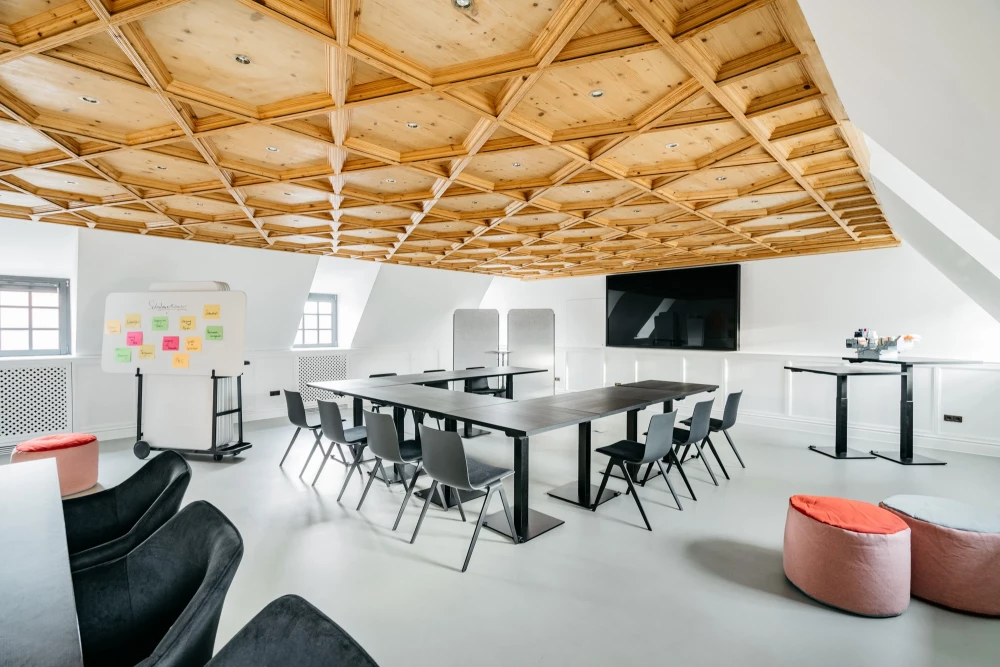 Moderner Konferenzraum mit Holzdeckenbalken und weißen Wänden, ausgestattet mit schwarzen Konferenzstühlen, einem großen Meetingtisch, Flipchart und Fernsehbildschirm. Zwei rote Sitzpoufs und ein schwarzer Sessel vervollständigen die minimalistische Einrichtung.