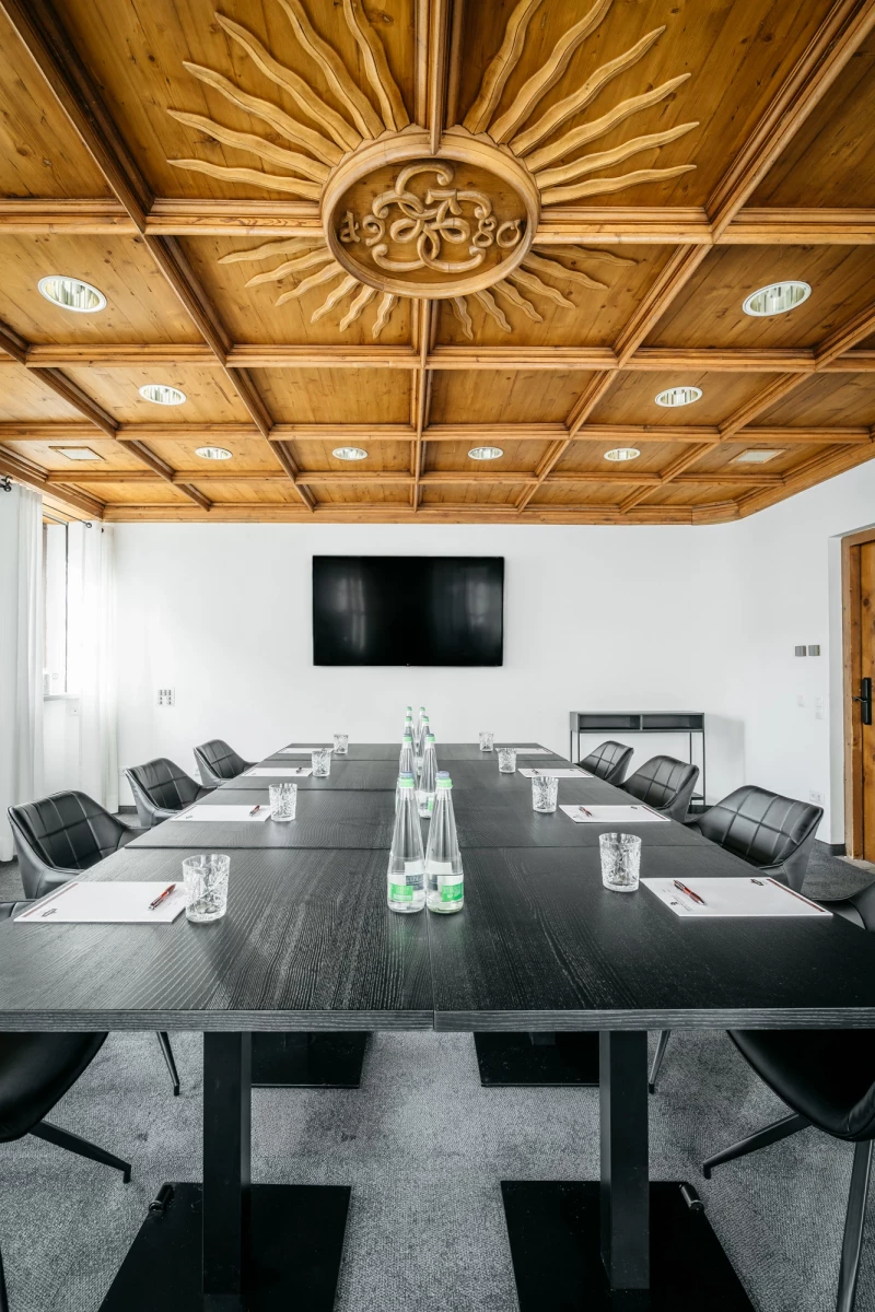 Moderner Konferenzraum mit schwarzem Konferenztisch, grauen Lederstühlen und einer Holzbalkendecke mit einem sonnenstrahlenförmigen Zentralmotiv. An der Wand hängt ein großer schwarzer Bildschirm, und auf dem Tisch stehen Wassergläser und Notizblöcke für Besprechungen.