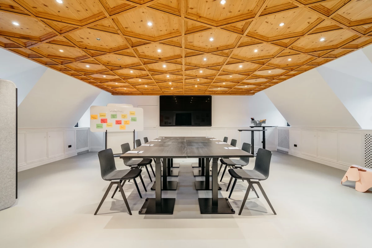 Moderner Konferenzraum mit Holzdecke und geometrischem Muster, ausgestattet mit einem großen Tisch, grauen Stühlen und einem schwarzen Bildschirm an der Wand. Helle, minimalistisch gestaltete Räumlichkeit mit indirekter Beleuchtung und einer Pinnwand mit bunten Haftnotizen.