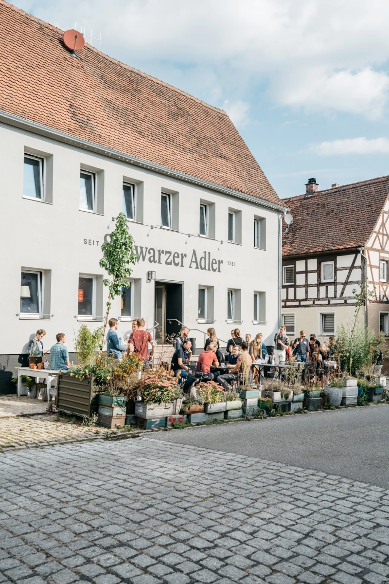 Straßenszene vor dem Gasthaus "Schwarzer Adler" mit Fachwerkhäusern, belebtem Außenbereich und Besuchern, die an Tischen und Pflanzkübeln vor dem weißen Gebäude sitzen.