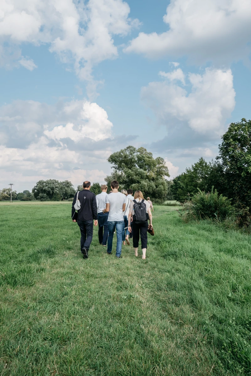 Fünf Personen gehen eine grüne Wiese entlang, im Hintergrund Bäume und ein blauer Sommerhimmel mit weißen Wolken. Die Gruppe trägt Alltagskleidung und bewegt sich von hinten gesehen.
