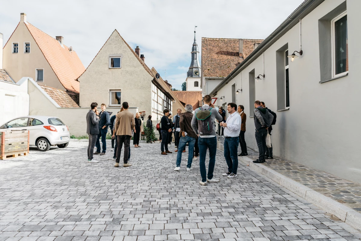 Eine Gruppe von Menschen steht auf einem gepflasterten Platz mit historischen Gebäuden und einem Kirchturm im Hintergrund. Die Teilnehmer sind in Grüppchen verteilt und tragen legere Kleidung, während sie sich offenbar im Gespräch austauschen.