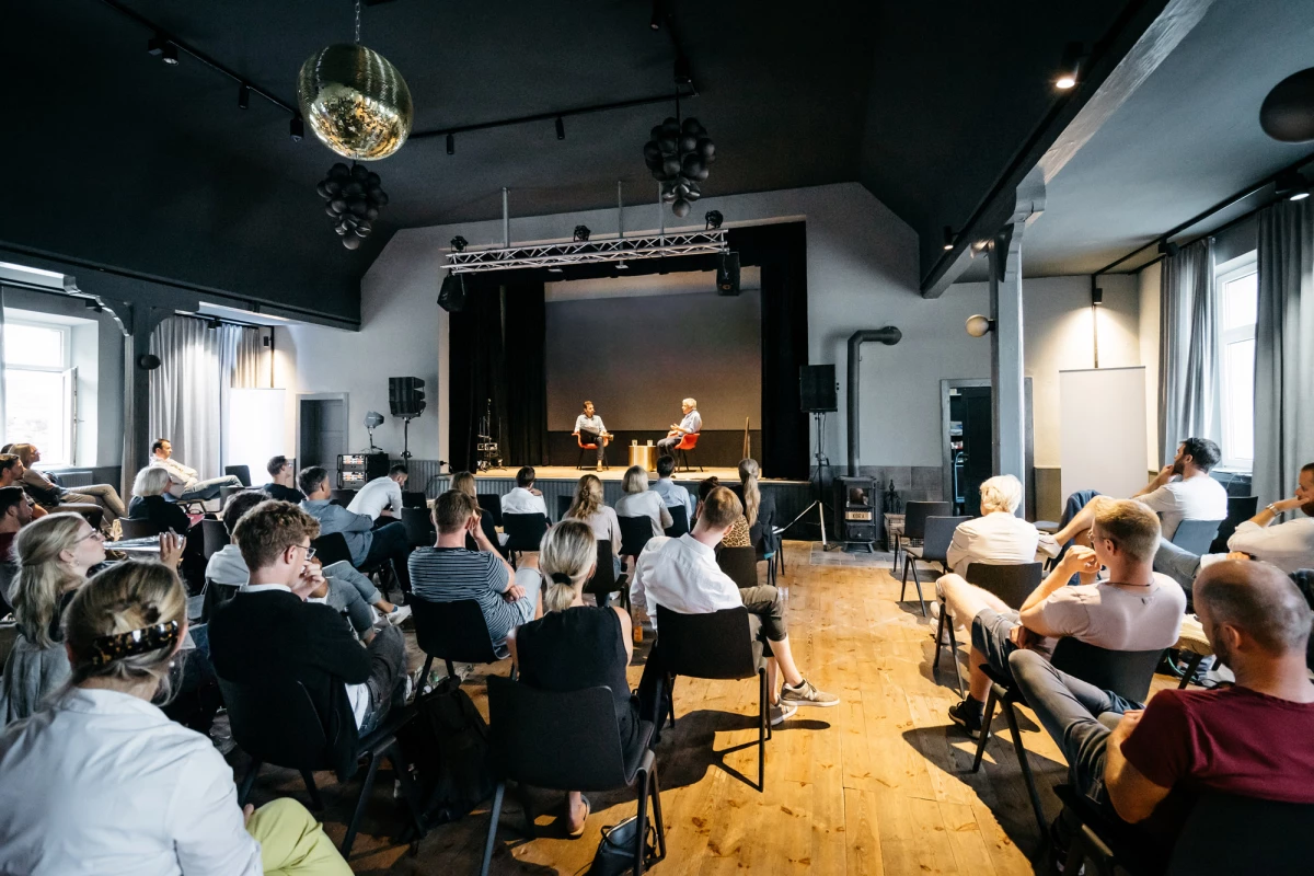 Zwei Personen sitzen auf einer Bühne vor einem Publikum, das aufmerksam einer Präsentation oder Diskussion in einem hell erleuchteten Veranstaltungsraum mit Holzboden und Discokugel folgt.