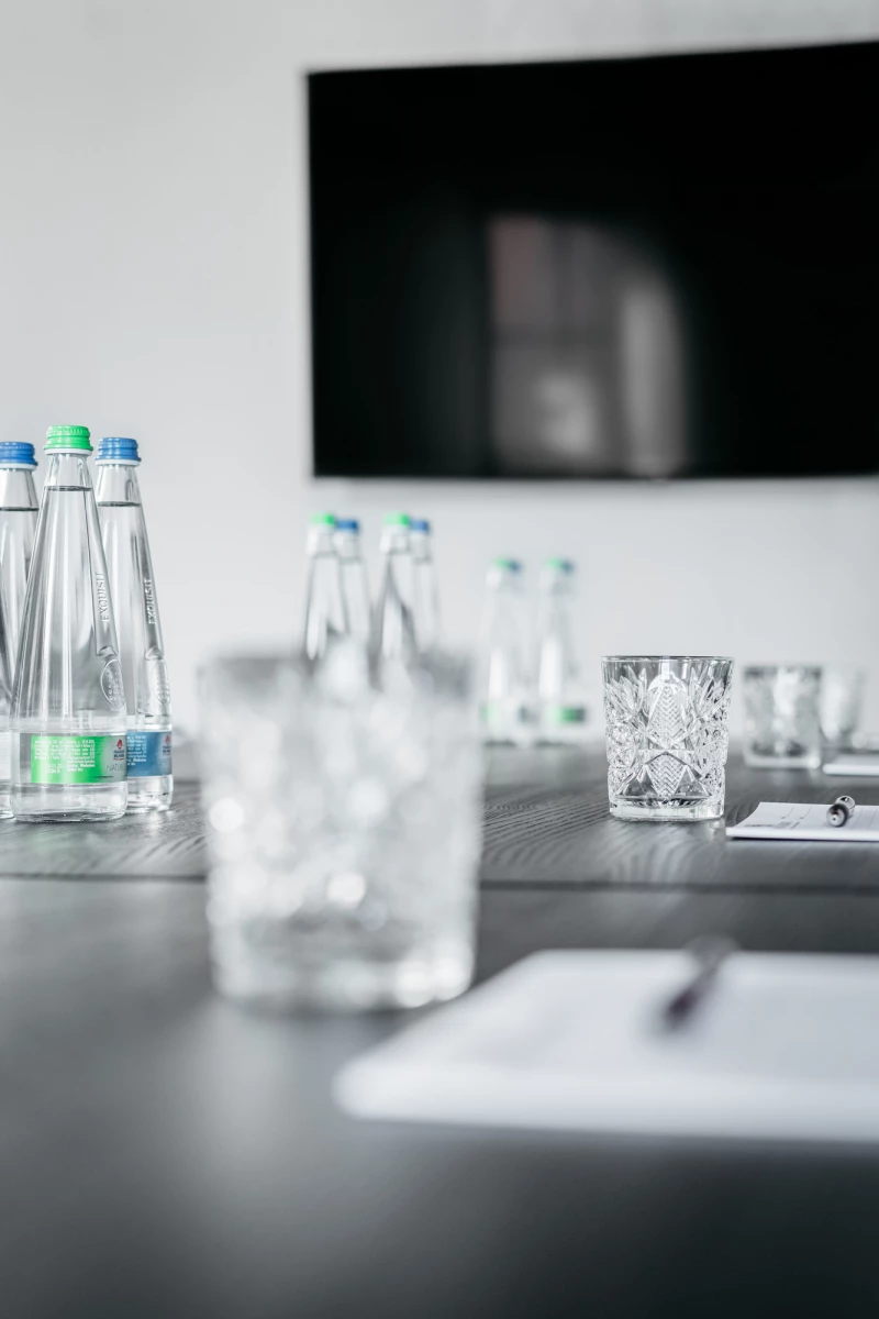 Glasflaschen mit Wasser und dekorativ geschliffene Wassergläser auf einem dunklen Tisch mit unscharfem Hintergrund, der einen Teil eines schwarzen Bildschirms oder Fernsehers zeigt.