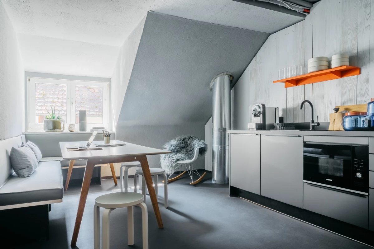 Moderne Dachgeschoss-Küche mit weißen Schränken, einem orangefarbenen Regal und einem hellen Holztisch mit Stühlen. Graue Wände, bodentiefe Fenster und eine minimalistische Einrichtung prägen den zeitgenössischen Wohnraum.