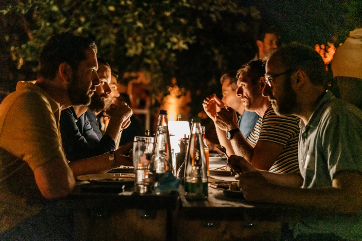 Abendliche Tischszene mit Personen, die an einem Tisch sitzen und miteinander in gedämpftem Licht kommunizieren, mit Getränken und einer intim-geselligen Atmosphäre.