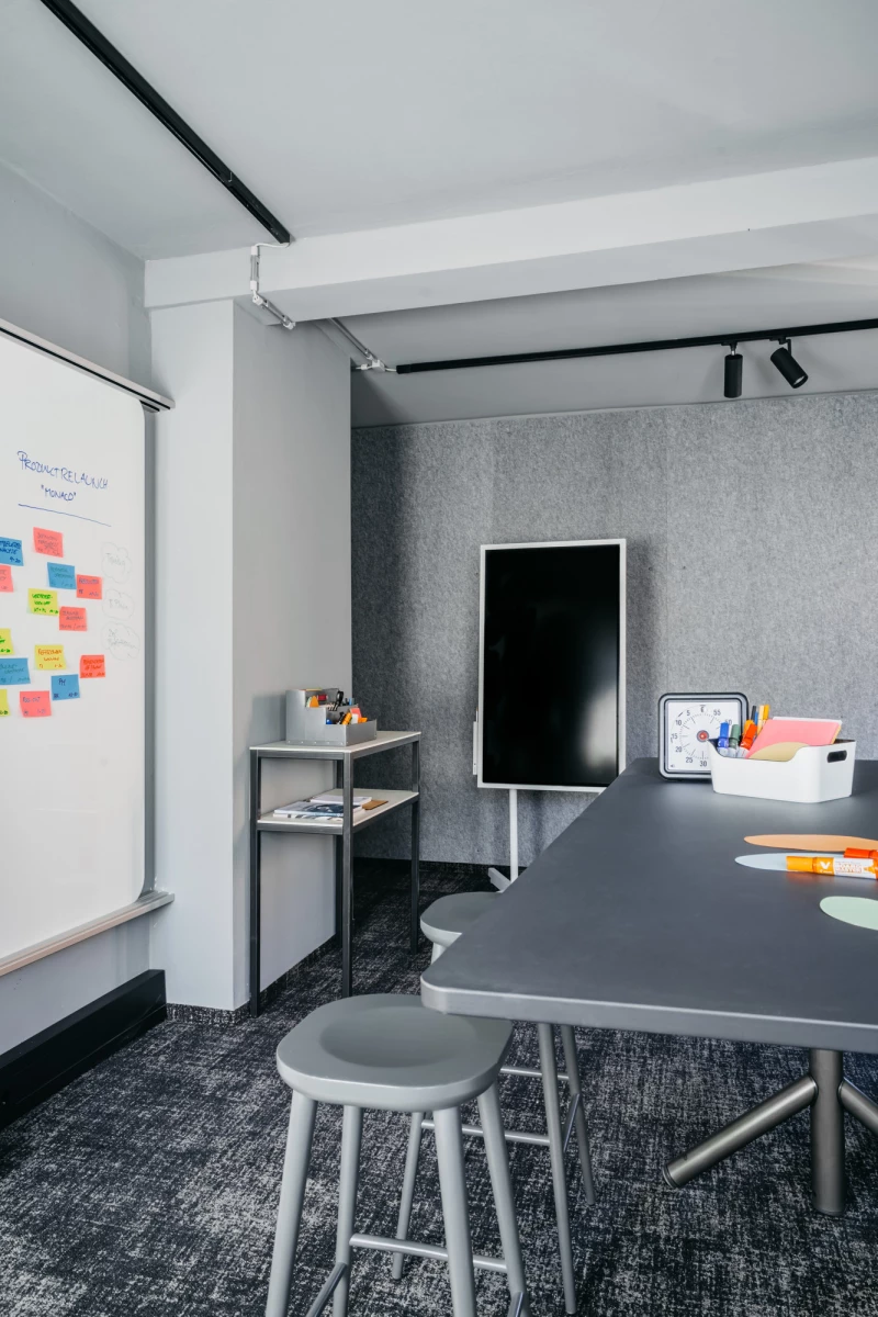 Moderner Besprechungsraum mit weißer Wandtafel, Post-its, grauem Tisch, Hocker und schwarzer Rückwand. Schwarze Deckenleuchten und Regale mit Utensilien vervollständigen das minimalistische Büroambiente.