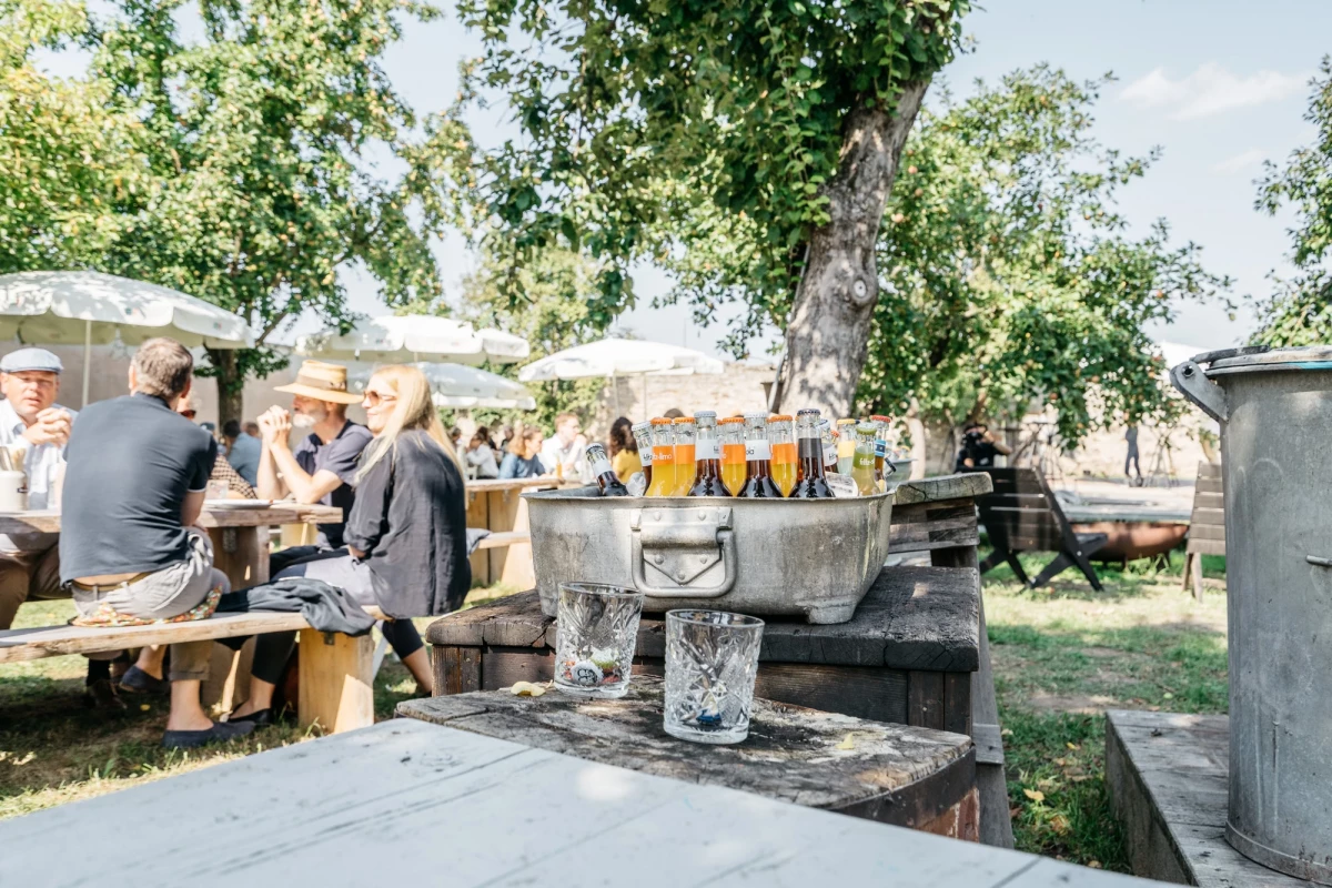 Biergläser stehen auf einem Steinpult in einem Biergarten mit Bäumen und sitzenden Personen an Holztischen im Hintergrund. Eine sommerliche Atmosphäre bei einem Treffen im Freien.