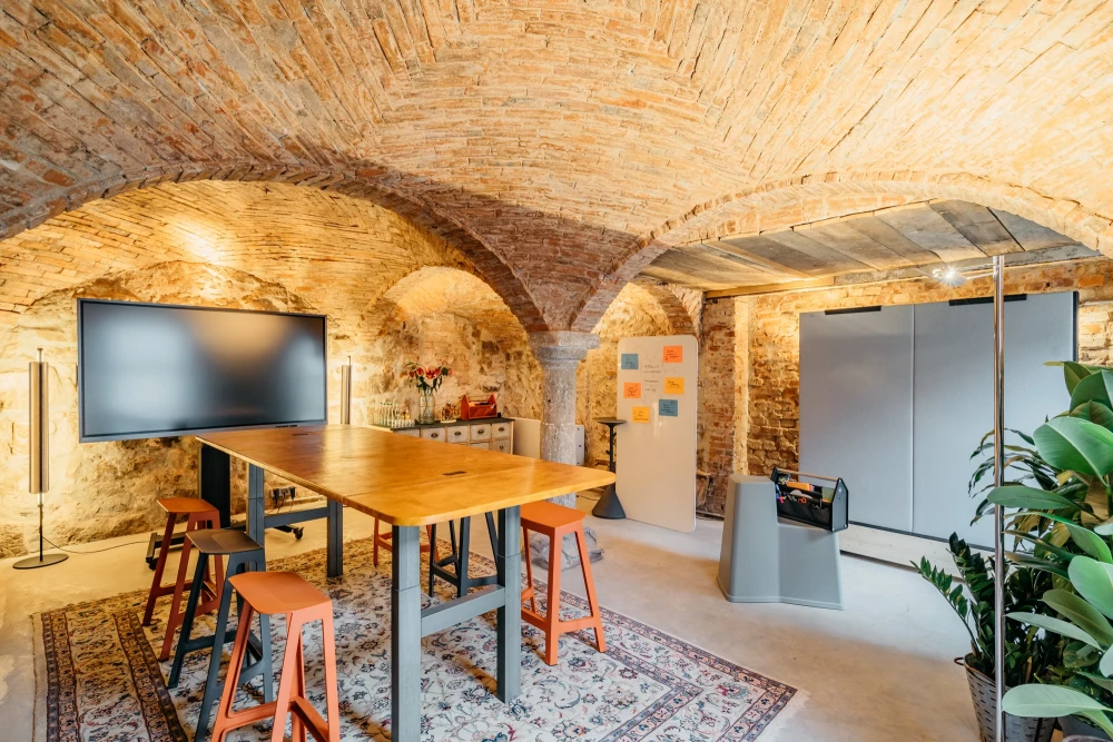 Moderner Besprechungsraum mit Backsteingewölbe und historischer Architektur. Ein großer Holztisch mit Barhockern steht auf einem gemusterten Teppich, daneben ein Flachbildschirm und eine Glastrennwand. Warmes Licht und rustikale Steinwände schaffen eine einladende Atmosphäre für Meetings.