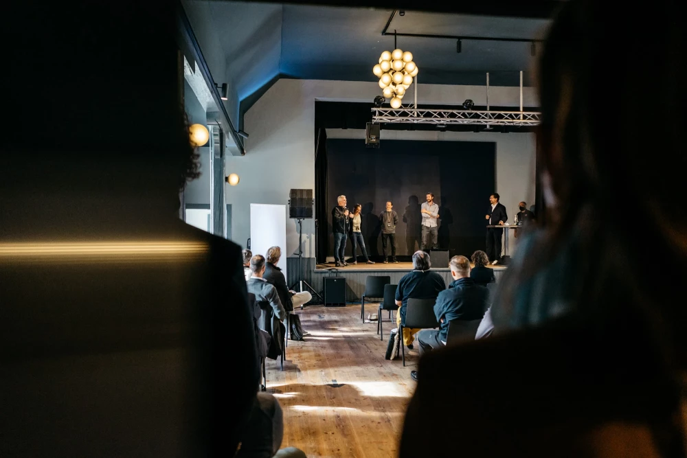 Mehrere Personen stehen auf einer Bühne vor einem Publikum in einem Veranstaltungsraum mit Holzboden und elegantem Kronleuchter. Die Atmosphäre wirkt professionell und konzentriert, mit Zuhörern, die der Präsentation aufmerksam folgen.