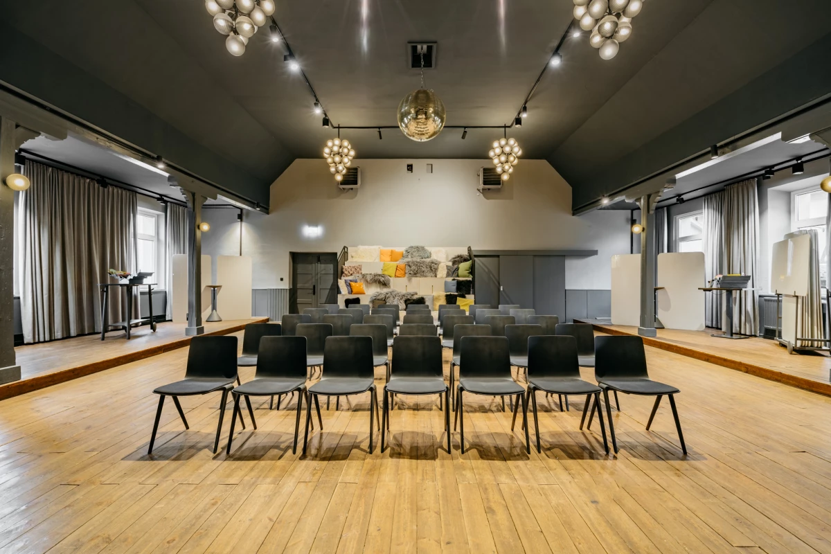 Moderner Konferenzraum mit hölzernem Fußboden, grauen Wänden und schwarzen Stühlen, die in Reihen aufgestellt sind. Große Deckenleuchten und Vorhänge an den Seitenwänden sorgen für eine einladende Atmosphäre. Ein Kunstwerk in gelb-grauen Tönen schmückt die Rückwand des Raumes.
