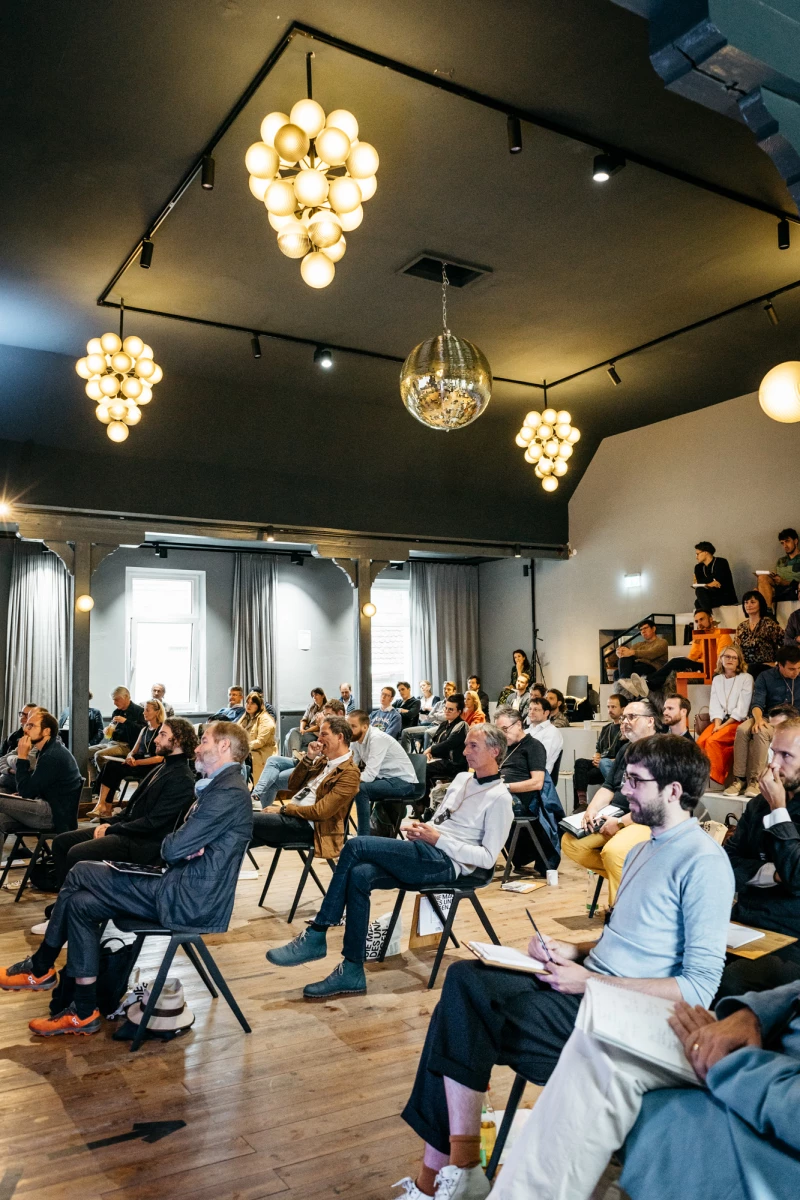 Teilnehmer einer Konferenz sitzen auf Stühlen in einem modern eingerichteten Raum mit auffälligen Deckenleuchten. Der Raum wirkt hell und einladend, mit Holzboden und großen Fenstern, während die Anwesenden aufmerksam einer Präsentation oder Diskussion folgen.