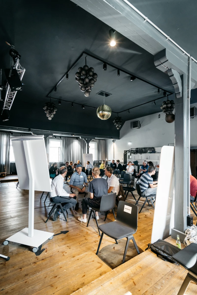 Seminarraum mit hoher Decke und Holzboden, in dem mehrere Personen an Tischen sitzen und an einem Workshop oder einer Konferenz teilnehmen. Moderne Beleuchtung und weiße Stellwände prägen den hellen, industriell anmutenden Raum.
