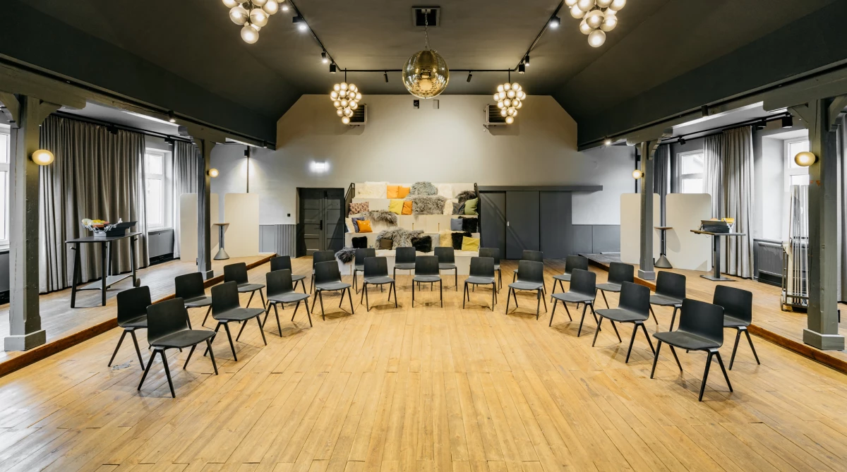 Heller Seminarraum mit Holzboden und modernen schwarzen Stühlen, die in einem Halbkreis angeordnet sind. Warme Beleuchtung durch Kugelleuchten und ein abstraktes Kunstwerk an der Wand im Hintergrund. Die Atmosphäre wirkt einladend und professionell für Meetings oder Workshops.