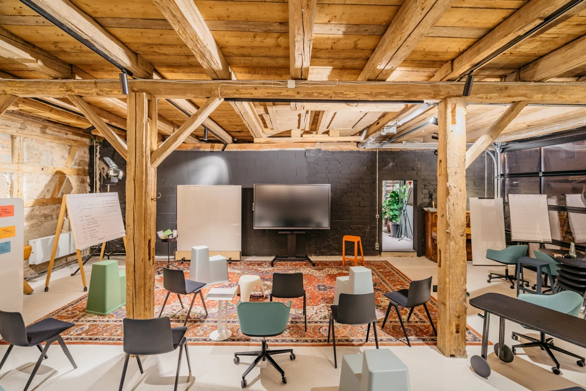 Moderner Besprechungsraum in einer Scheune mit Holzbalkendecke, ausgestattet mit schwarzen Drehstühlen, einem großen Fernseher und einem bunten Teppich. Durchdachtes Industriedesign mit natürlichen Holzelementen schafft eine inspirierende Arbeitsatmosphäre.