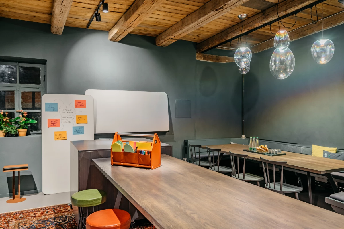 Moderne Teamküche mit Holzdeckenbalken, grauen Wänden und hängenden Glaslampen. Ein langer Holztisch mit orangefarbener Werkzeugkiste und bunten Hockern, daneben eine Whiteboard-Wand mit Haftnotizen. Große Fenster und eine gemütliche Arbeitsatmosphäre prägen den Raum.