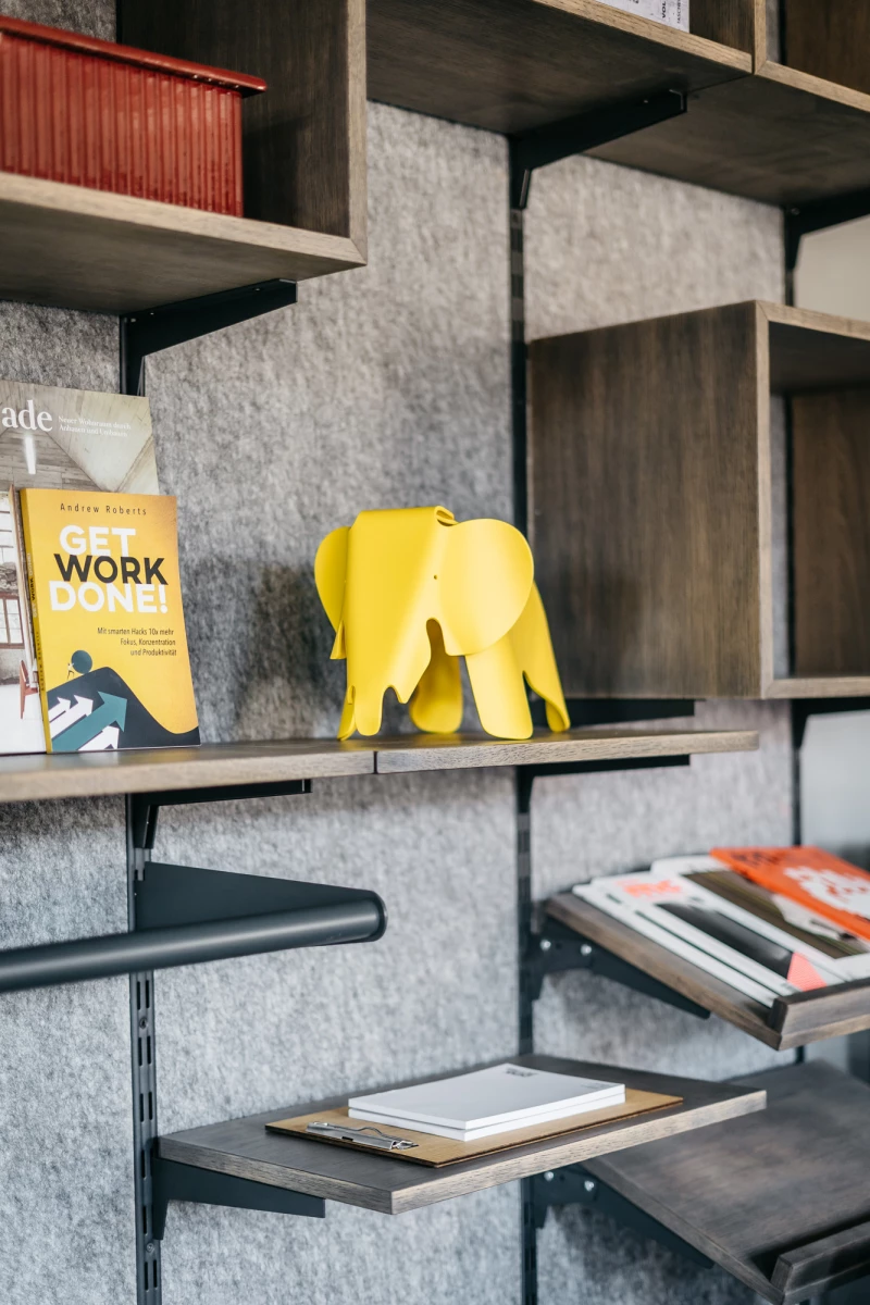 Graues Bücherregal mit gelber Elefantenfigur und Büchern, darunter ein gelbes Buch mit dem Titel "Work done". Die Regalbretter sind aus Holz und mit Filz ausgelegt, was eine gemütliche und moderne Atmosphäre schafft.