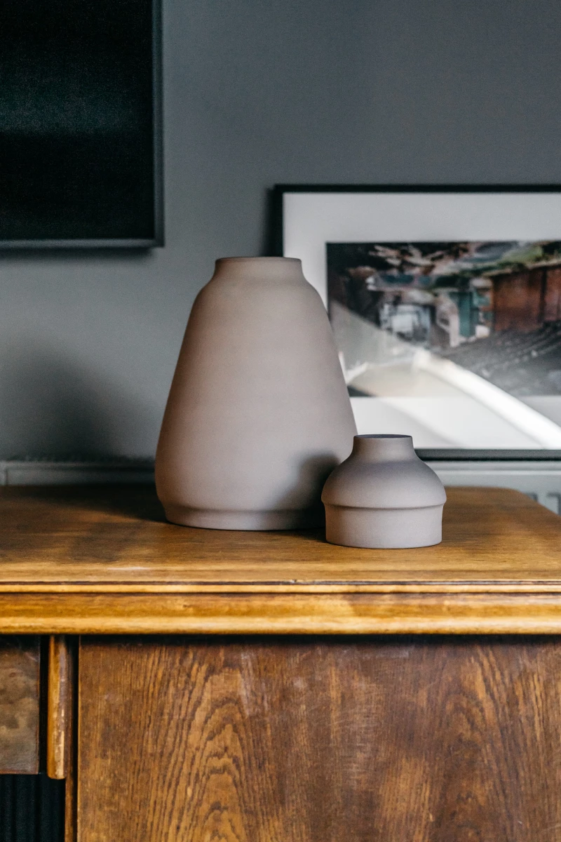 Zwei beige Vasen unterschiedlicher Größe stehen auf einem Holzregal vor einem gerahmten Kunstdruck. Die Vasen haben eine matte, glatte Oberfläche und fügen sich harmonisch in das minimalistisch-skandinavische Wohnambiente ein.
