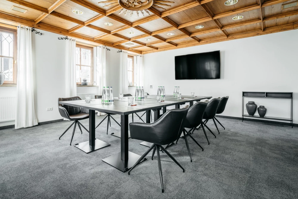 Moderner Konferenzraum mit weißen Wänden, holzgetäfelter Decke und grauem Teppichboden. Ein großer rechteckischer Konferenztisch mit schwarzen Designerstühlen steht im Raum, an der Wand hängt ein großer schwarzer Fernseher. Die Fenster sind mit weißen Vorhängen verhangen.