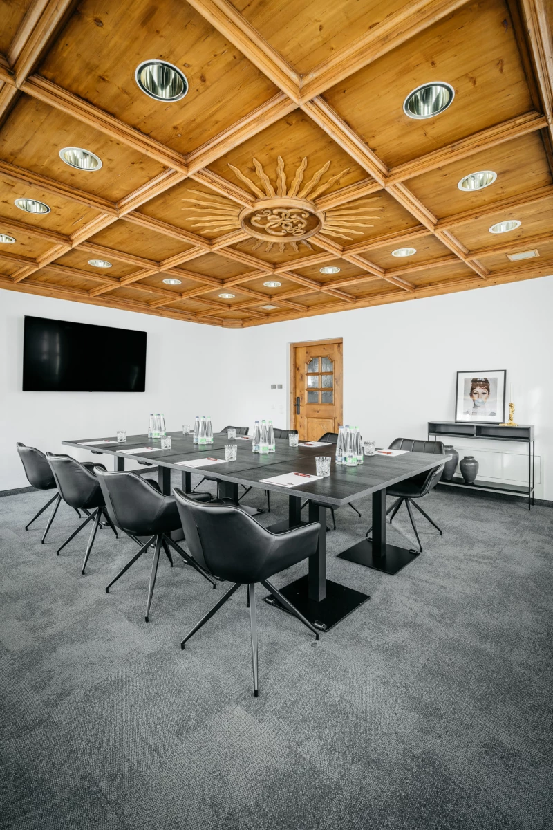 Moderner Konferenzraum mit Holzdeckengestaltung, großem Glastisch und schwarzen Drehstühlen. Schwarze Wandtafel und graue Teppichbodengestaltung vervollständigen den eleganten Raum.