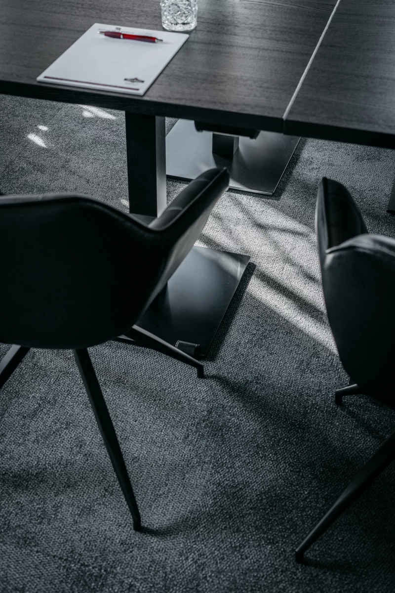 Moderner Konferenztisch mit Papier und Stift, umgeben von grauen Designerstühlen auf einem dunkleren Teppichboden. Scharfe Lichtschatten fallen diagonal über die Szenerie und betonen die klare, geometrische Komposition des Raumes.