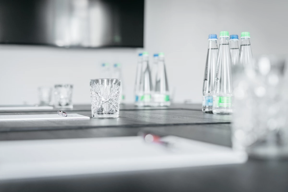 Transparente Wassergläser und Glasflaschen stehen auf einem hellen Tisch oder Thekenbereich. Die Komposition wirkt minimalistisch und elegant, mit einem unscharfen Hintergrund in gedämpften Grautönen.