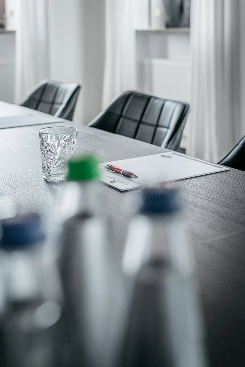 Holzerner Konferenztisch mit einem geschliffenen Wasserglas und einem Notizbuch, umgeben von schwarzen Lederstühlen in einem hellen, minimalistisch eingerichteten Besprechungsraum.