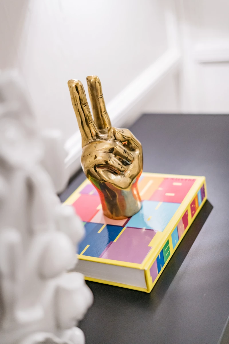 Goldene Hand-Skulptur mit Victory-Zeichen, die auf einem bunten, mehrfarbigen Buch oder Kasten mit gelben Kanten sitzt. Die Skulptur ist auf einer dunklen Oberfläche vor einem hellen Hintergrund platziert.