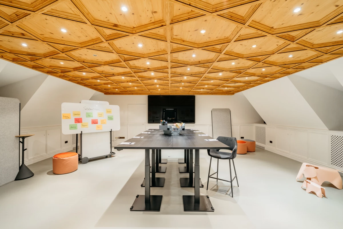 Moderner Besprechungsraum mit geometrischer Holzdecke und Designerbeleuchtung. Ein großer Konferenztisch mit Stühlen steht in der Mitte, daneben eine Pinnwand und ein Whiteboard. Der Raum wirkt hell, minimalistisch und einladend, mit weißen Wänden und warmen Holztönen.