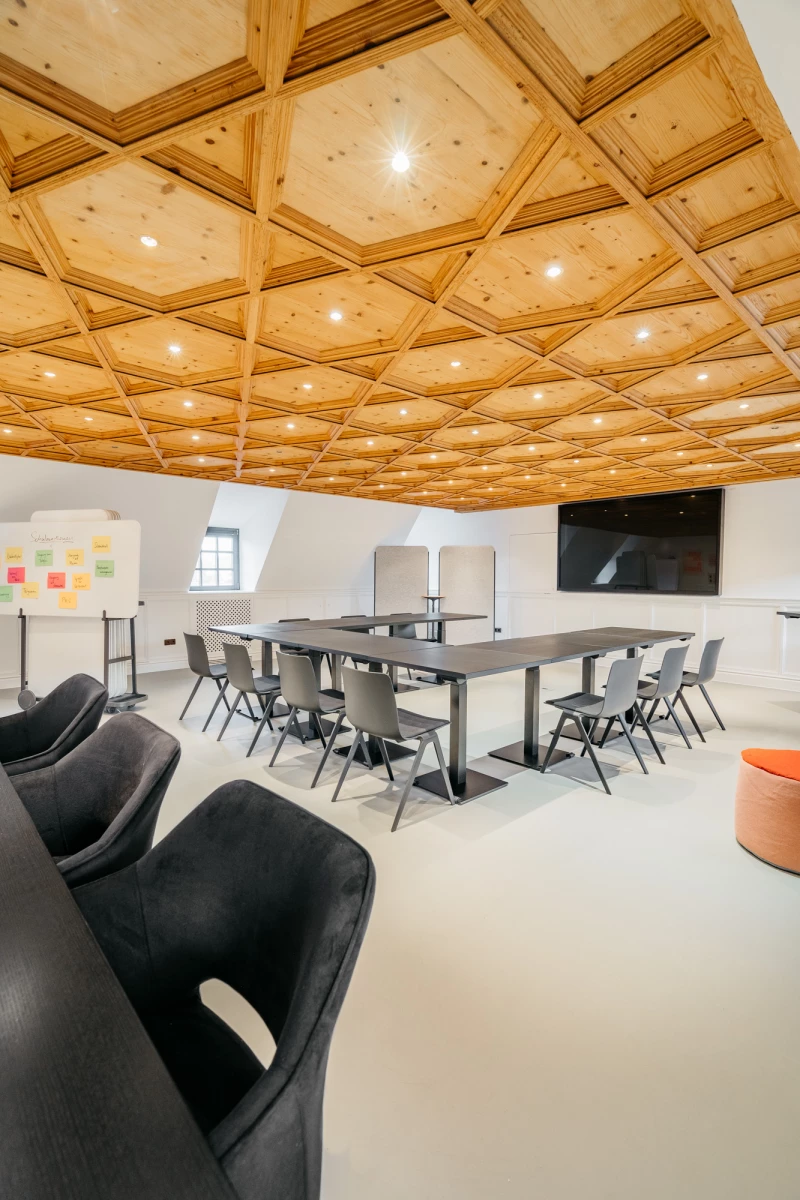 Moderner Konferenzraum mit holzgetäfelter Decke und elegant designten Möbeln. Graue Tische und Stühle stehen in U-Form, mit schwarzen Sesseln im Vordergrund und einer weißen Wand mit bunten Haftnotizen. Warmes Licht und große Fenster sorgen für eine einladende Arbeitsatmosphäre.