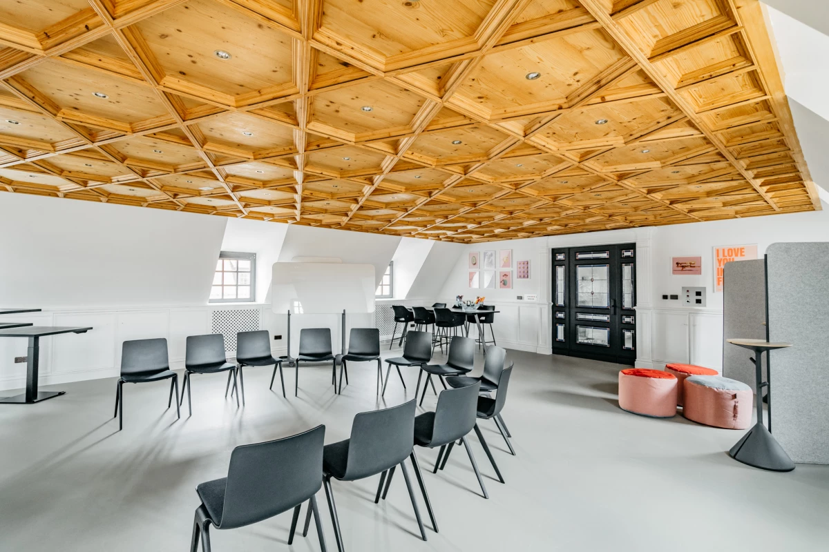 Heller Seminarraum mit holzgetäfelter Decke und modernen grauen Stühlen, die in Reihen angeordnet sind. Zwei Fenster und eine schwarze Tür an der Wand, daneben rosa Sitzpuffs und technische Geräte.