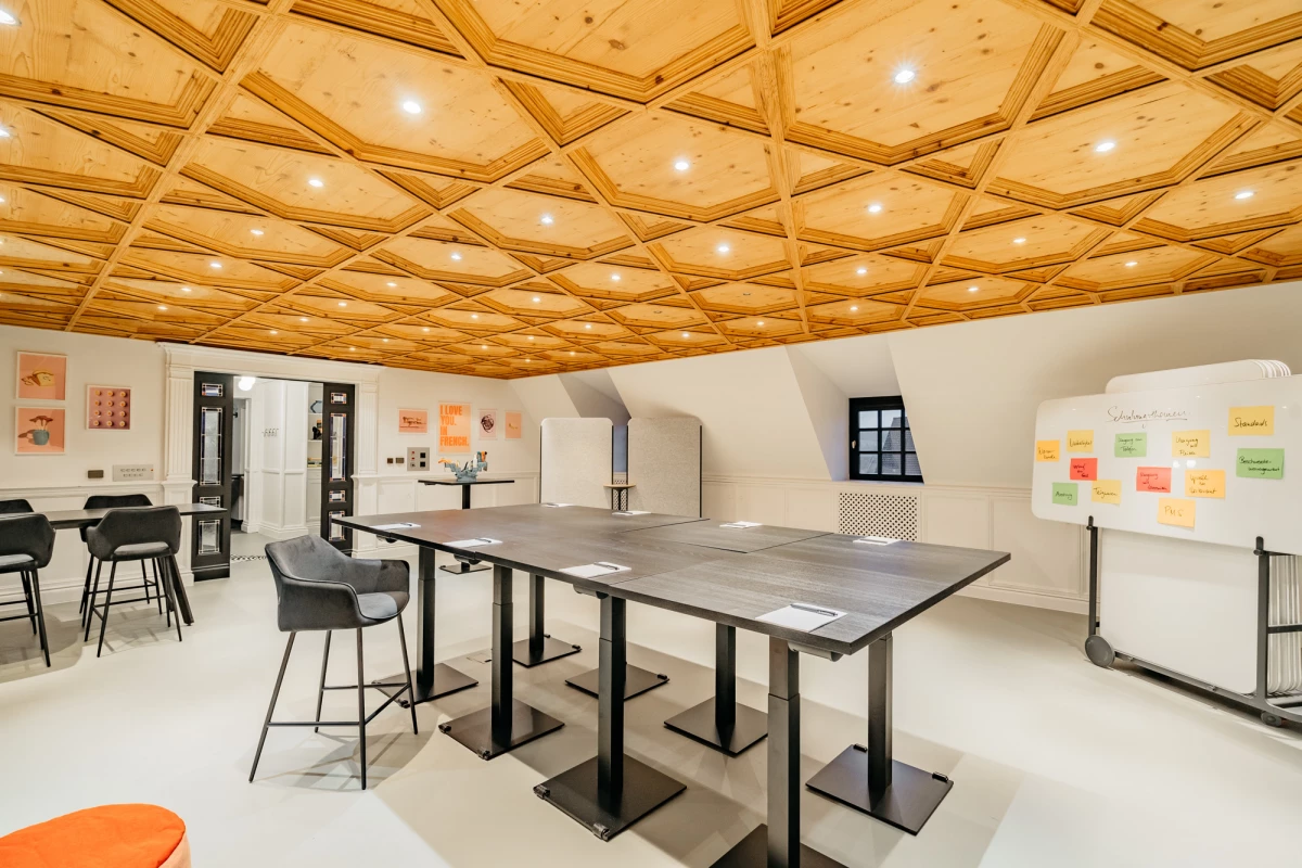 Moderner Besprechungsraum mit elegantem Holzdeckendesign, grauen Arbeitstischen und schwarzen Stühlen. Helle, offene Atmosphäre mit eingebauten Deckenleuchten und einer Pinnwand an der Wand. Professioneller Arbeitsbereich mit natürlichem Licht und zeitgemäßem Mobiliar.