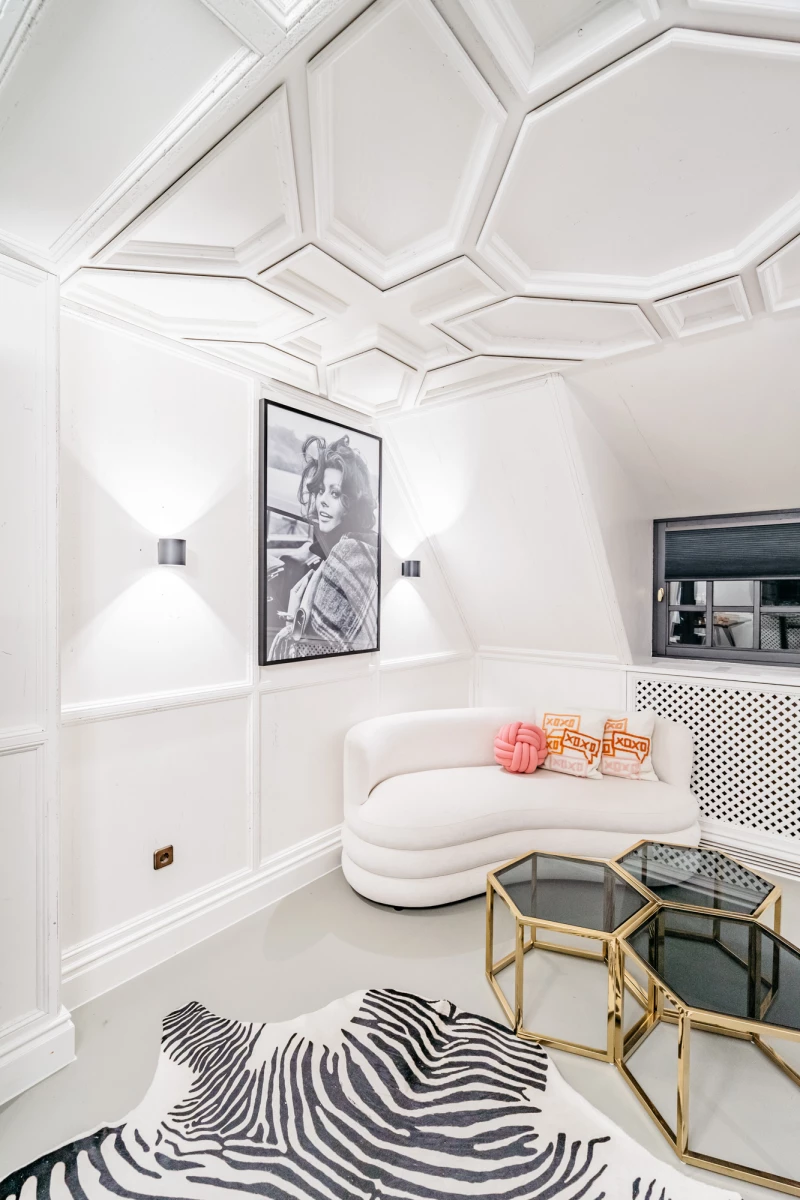 Moderner, weißer Wohnraum mit geschwungener Couch und geometrischer Deckengestaltung. Schwarzweißes Kunstwerk an der Wand, Zebra-gemusterter Teppich und goldene Beistelltische bilden einen eleganten, minimalistischen Raum.