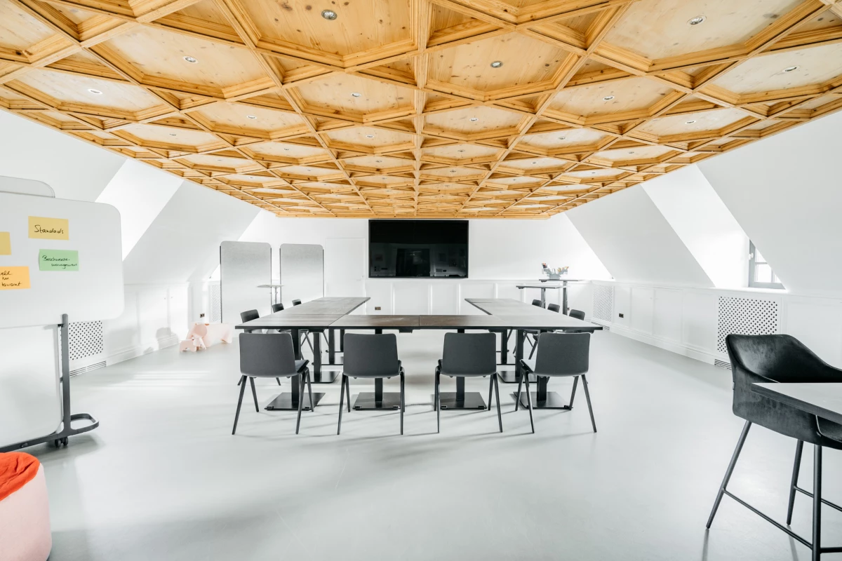 Moderner Besprechungsraum mit geometrischer Holzdecke, weißen Wänden und schwarzen Stühlen um einen rechteckigen Tisch. Ein schwarzer Bildschirm hängt an der Wand, und der Raum wirkt hell und minimalistisch gestaltet.