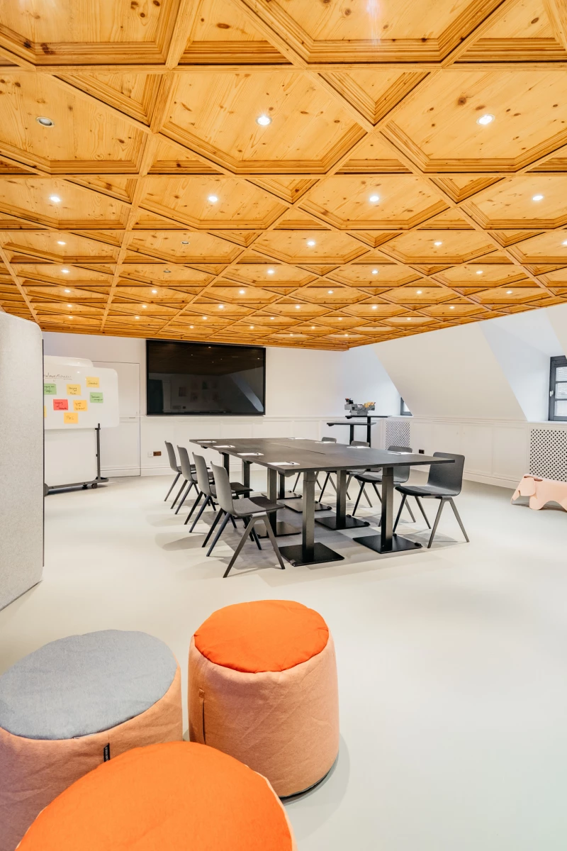 Moderner Konferenzraum mit Holzdecke und geometrischen Einbauleuchten, ausgestattet mit grauen Bürostühlen um einen Konferenztisch, ergänzt durch orange und graue Sitzpoufs im Vordergrund und einer Pinnwand an der Wand.