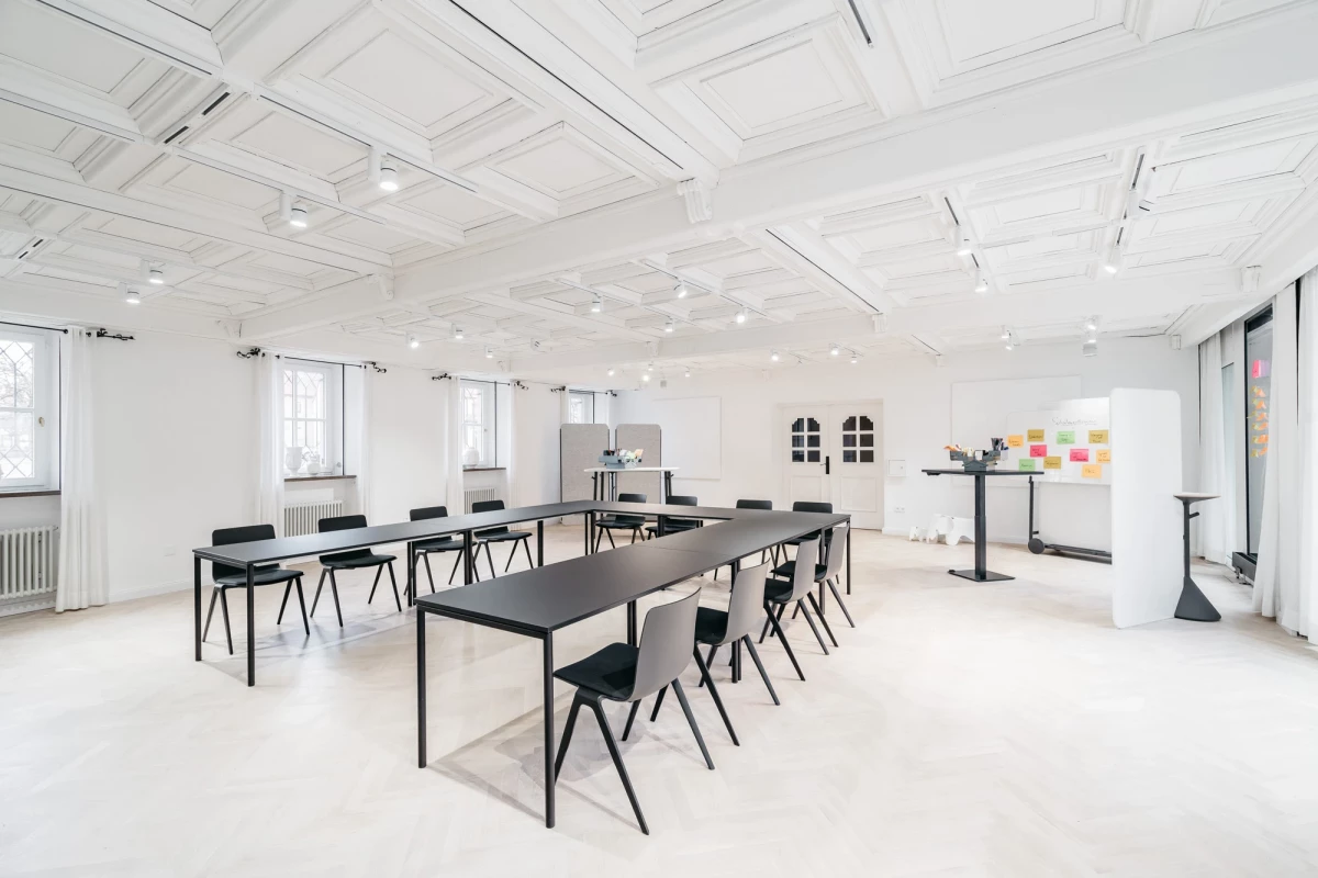 Heller, minimalistischer Seminarraum mit langen schwarzen Tischen und passenden Stühlen. Weiße Wände und Decke mit Oberlicht, große Fenster sorgen für natürliches Licht. An der Wand hängen bukte Haftnotizen, ein Whiteboard steht bereit für Besprechungen und Workshops.