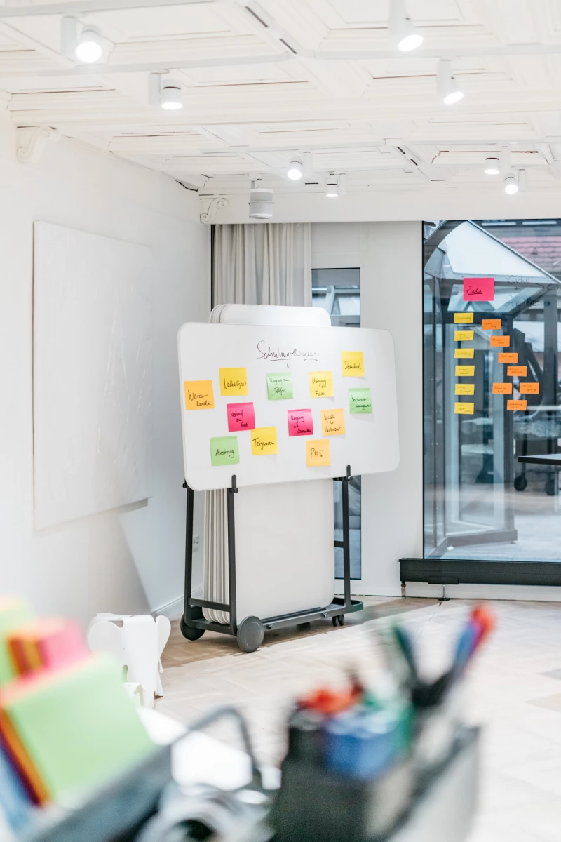 Whiteboard mit bunten Haftnotizen auf einem mobilen Ständer in einem hellen, modernen Büroraum mit Glaswand und Deckenspots. Verschiedene Farben der Notizen deuten auf eine Brainstorming- oder Planungssitzung hin.