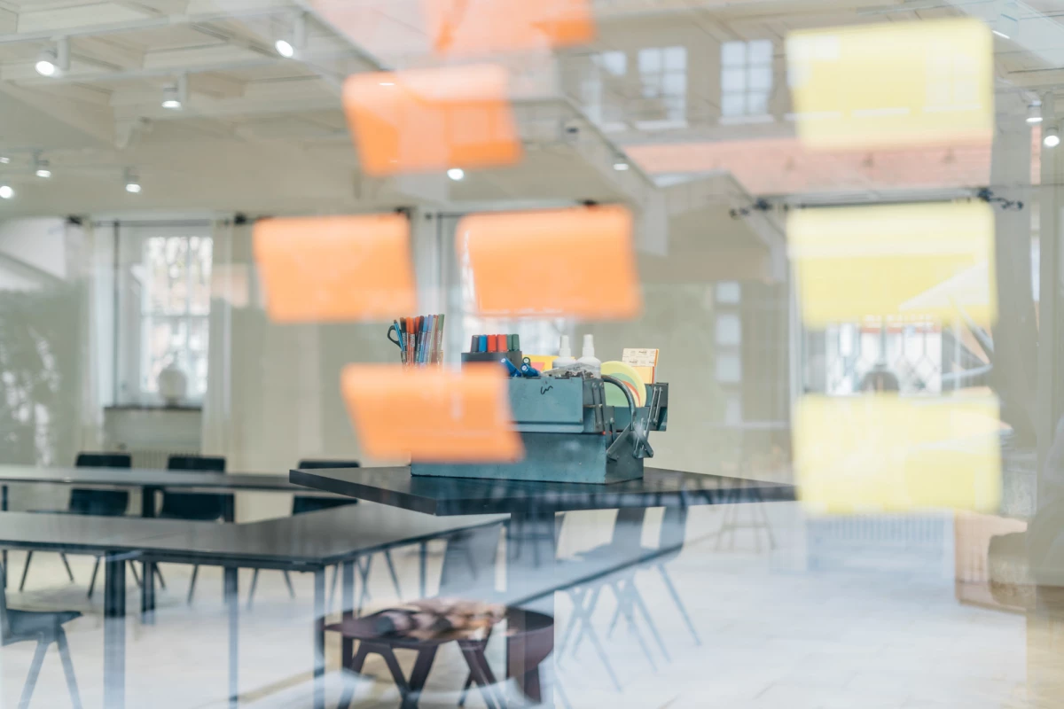 Blick in einen lichtdurchfluteten Besprechungsraum mit Glastisch und orangefarbenen Haftnotizen im Vordergrund. Moderne Büroeinrichtung mit Stühlen und gedämpftem Tageslicht, das durch große Fenster fällt.
