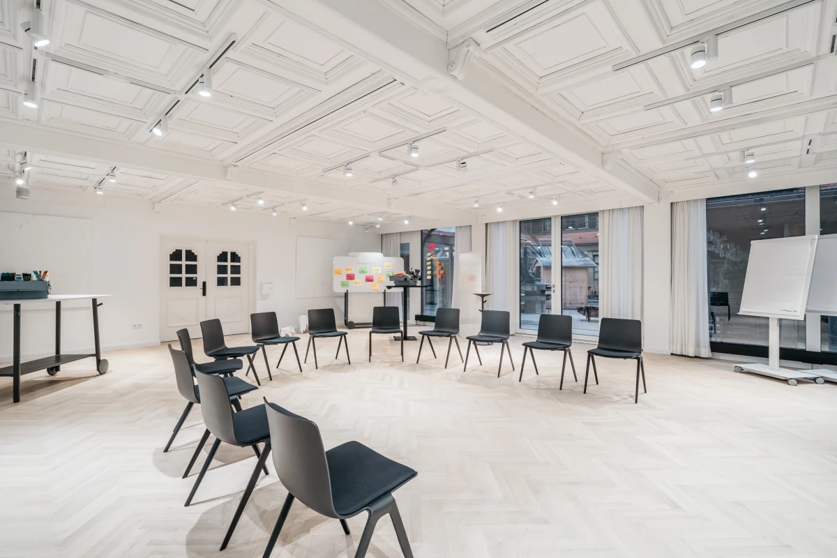 Heller, minimalistischer Seminarraum mit weißen Wänden und Decke, ausgestattet mit schwarzen Stühlen, die in einem Halbkreis angeordnet sind. Große Fenster lassen viel natürliches Licht herein, und Deckenspots sorgen für zusätzliche Beleuchtung. Ein Whiteboard an der Seite deutet auf einen Schulungs- oder Besprechungsraum hin.